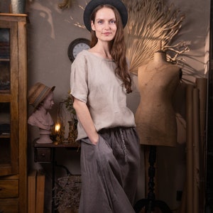 Model indoors wearing dark gray linen maxi skirt with elastic waistband, drawstring waist, and side pockets; loose, floor-length silhouette. Paired with light beige linen blouse and black wide-brim hat. Room includes vintage bust, clock, cabinet, and dried plants. Keywords: adjustable waist skirt, linen summer skirt.
