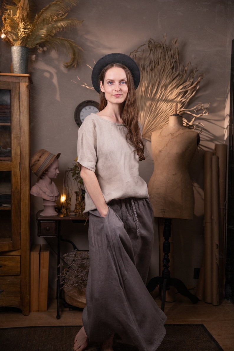 Model indoors wearing dark gray linen maxi skirt with elastic waistband, drawstring waist, and side pockets; loose, floor-length silhouette. Paired with light beige linen blouse and black wide-brim hat. Room includes vintage bust, clock, cabinet, and dried plants. Keywords: adjustable waist skirt, linen summer skirt.