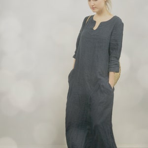 One maxi linen summer dress in deep navy with simple kaftan shape and pockets, worn by a model standing against a soft light background inside.