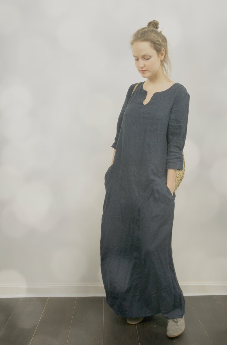 One maxi linen summer dress in deep navy with simple kaftan shape and pockets, worn by a model standing against a soft light background inside.