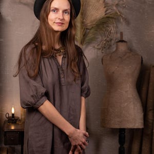 Dark gray-brown linen dress worn on a model, front three-quarter full-length crop, with round gathered neckline, short button placket, elbow-length puff sleeves, cuff bands, and loose straight shape.