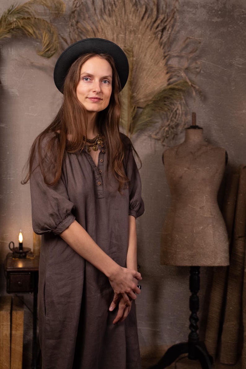 Dark gray-brown linen dress worn on a model, front three-quarter full-length crop, with round gathered neckline, short button placket, elbow-length puff sleeves, cuff bands, and loose straight shape.