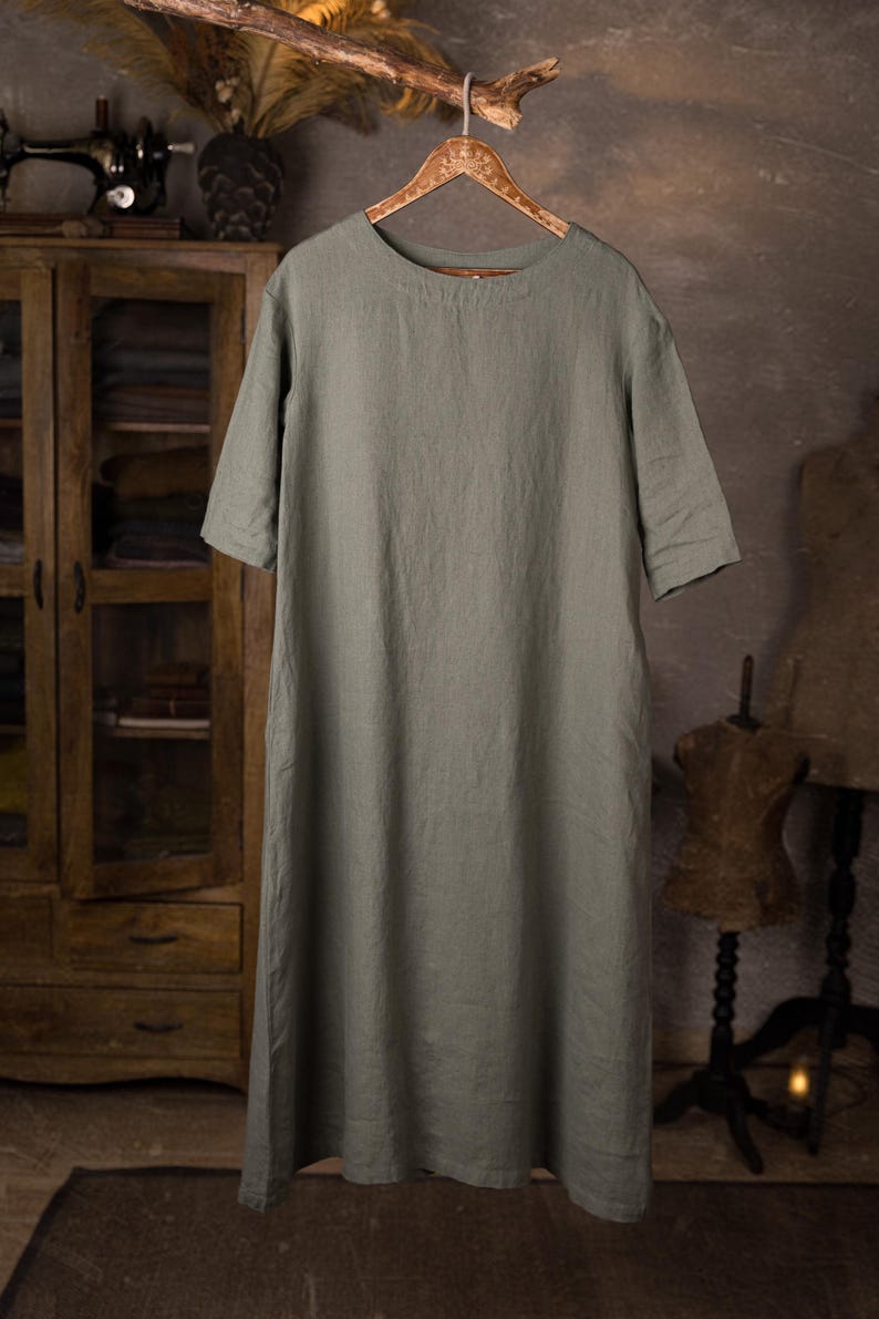 Full-length front view of sage linen dress hanging on a decorative wooden hanger, round neckline, short sleeves, relaxed drape with light wrinkles, plain front, rope loop over a wooden branch; blurred cabinet and mannequin in background, knockknocklinen.