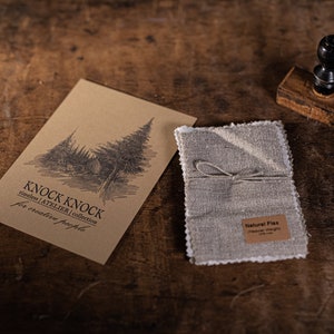Puede incluir: Una tarjeta de papel marrón con un dibujo de árboles y el texto "KNOCK KNOCK atelier | collection for creative people". Junto a la tarjeta hay una pequeña bolsa de lino atada con una cuerda y una etiqueta que dice "Natural Flax". También hay un sello de madera y unas tijeras sobre la superficie de madera.