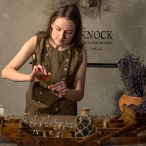 SPARROW linen vest in brown, displayed on a model in a front mid-length view, with a sleeveless shape, round neckline, double rows of light buttons, a slanted front edge, and a fitted body beside glass bottles and dried lavender, Knock Knock Linen.