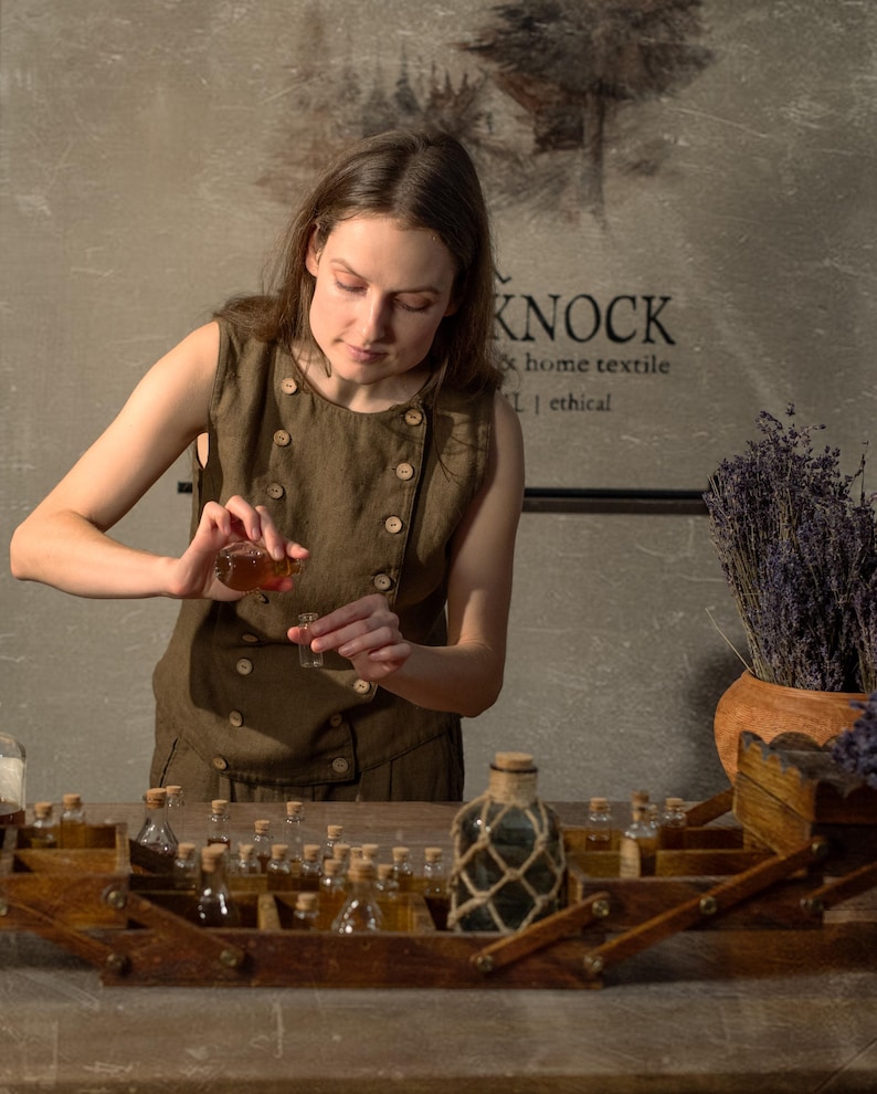 SPARROW linen vest in brown, displayed on a model in a front mid-length view, with a sleeveless shape, round neckline, double rows of light buttons, a slanted front edge, and a fitted body beside glass bottles and dried lavender, Knock Knock Linen.