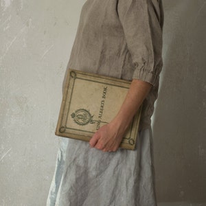 Person standing sideways against a textured wall, wearing a loose-fitting blouse made from natural flax linen. The top features a boat neckline and gathered cuffs, styled with a light grey skirt and vintage book. Linen blouse, boat neck top.
