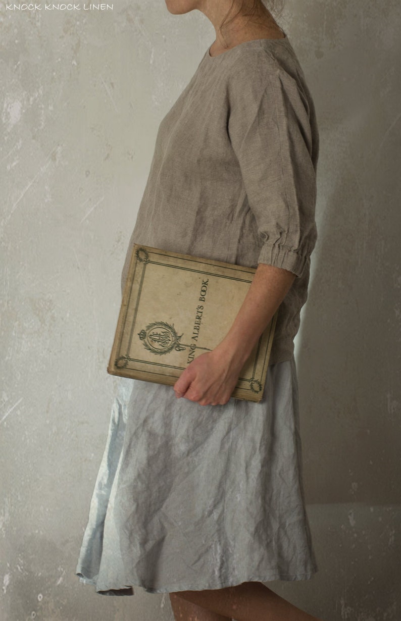 Person standing sideways against a textured wall, wearing a loose-fitting blouse made from natural flax linen. The top features a boat neckline and gathered cuffs, styled with a light grey skirt and vintage book. Linen blouse, boat neck top.