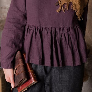 Detail view of a dark purple linen blouse with long sleeves and ruffled peplum waist, shown on a model standing indoors in grey checked trousers.