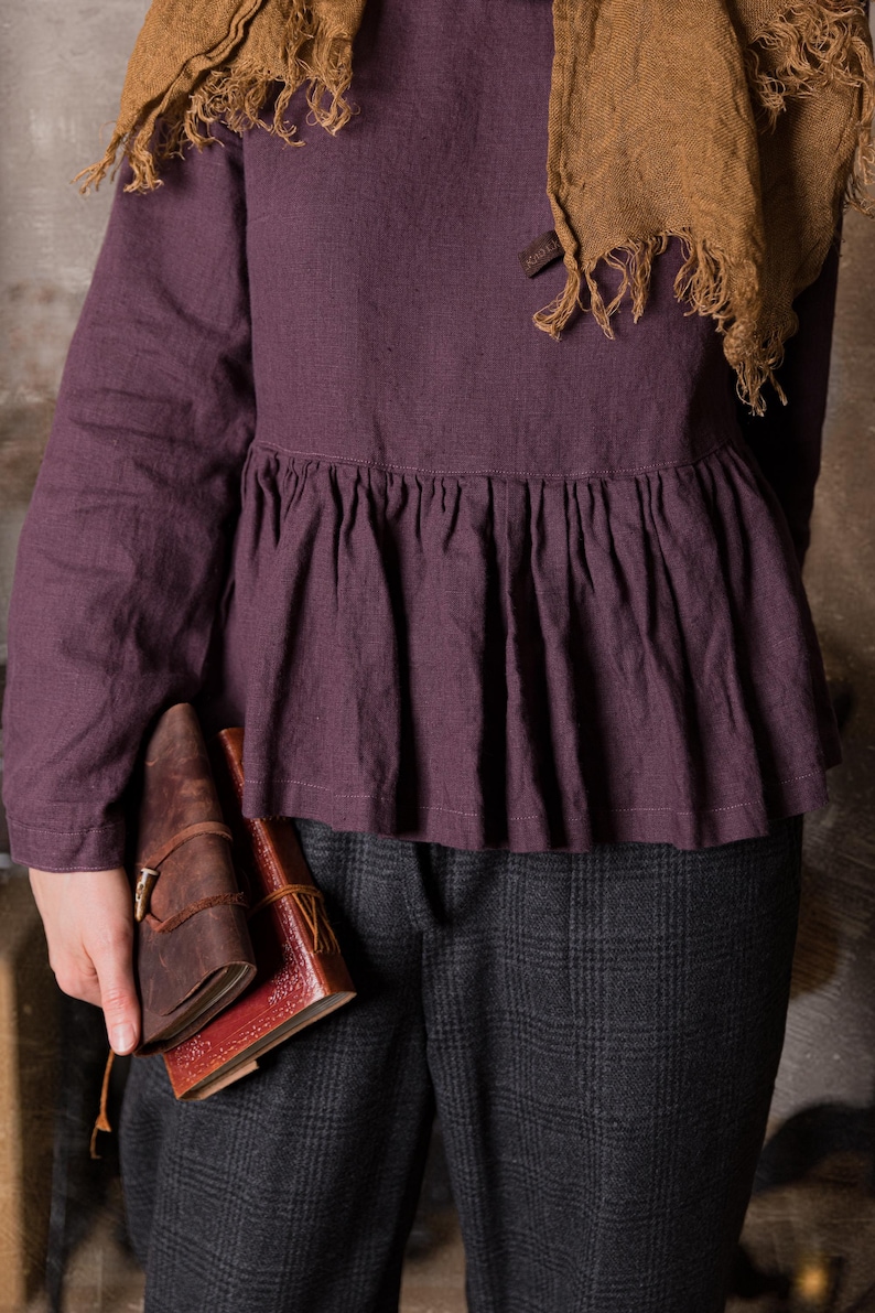 Detail view of a dark purple linen blouse with long sleeves and ruffled peplum waist, shown on a model standing indoors in grey checked trousers.