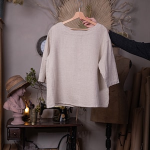 May include: A beige linen top with a boat neck and three-quarter sleeves hangs on a wooden hanger. The top is on a mannequin in a room with a sewing machine and other crafting supplies.
