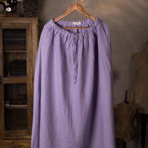 Lavender linen maxi skirt shown full-length on an ornate wooden hanger, front view, elasticated gathered waist, drawstring channel with hanging ties, wide A-line shape, curved hemline, cabinet and mannequin forms in the background.