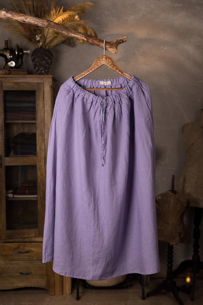 Lavender linen maxi skirt shown full-length on an ornate wooden hanger, front view, elasticated gathered waist, drawstring channel with hanging ties, wide A-line shape, curved hemline, cabinet and mannequin forms in the background.