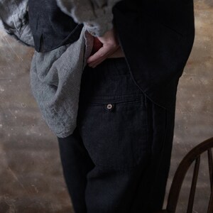 Linen pants in dark charcoal, shown on a model in a close-up side detail view, with a back patch pocket, small button on the pocket welt, straight seam lines, a loose leg, and a gray striped shirt draped over the hip, Knock Knock Linen.