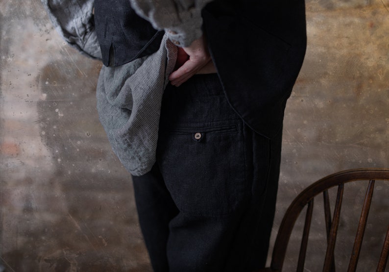 Linen pants in dark charcoal, shown on a model in a close-up side detail view, with a back patch pocket, small button on the pocket welt, straight seam lines, a loose leg, and a gray striped shirt draped over the hip, Knock Knock Linen.
