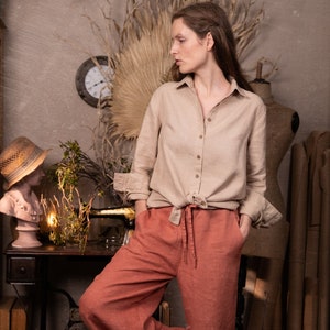 Dusty clay linen palazzo pants WIND on model, full-body three-quarter view; drawstring tie at center waist, hands in side pockets, wide legs with soft creases, one knee bent, beige button shirt knotted at waist, studio props behind.
