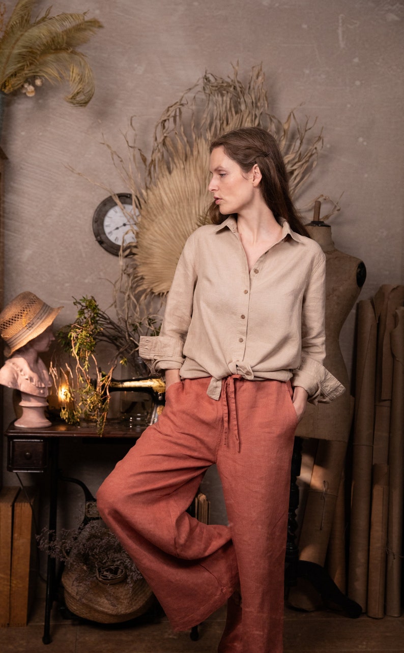Dusty clay linen palazzo pants WIND on model, full-body three-quarter view; drawstring tie at center waist, hands in side pockets, wide legs with soft creases, one knee bent, beige button shirt knotted at waist, studio props behind.