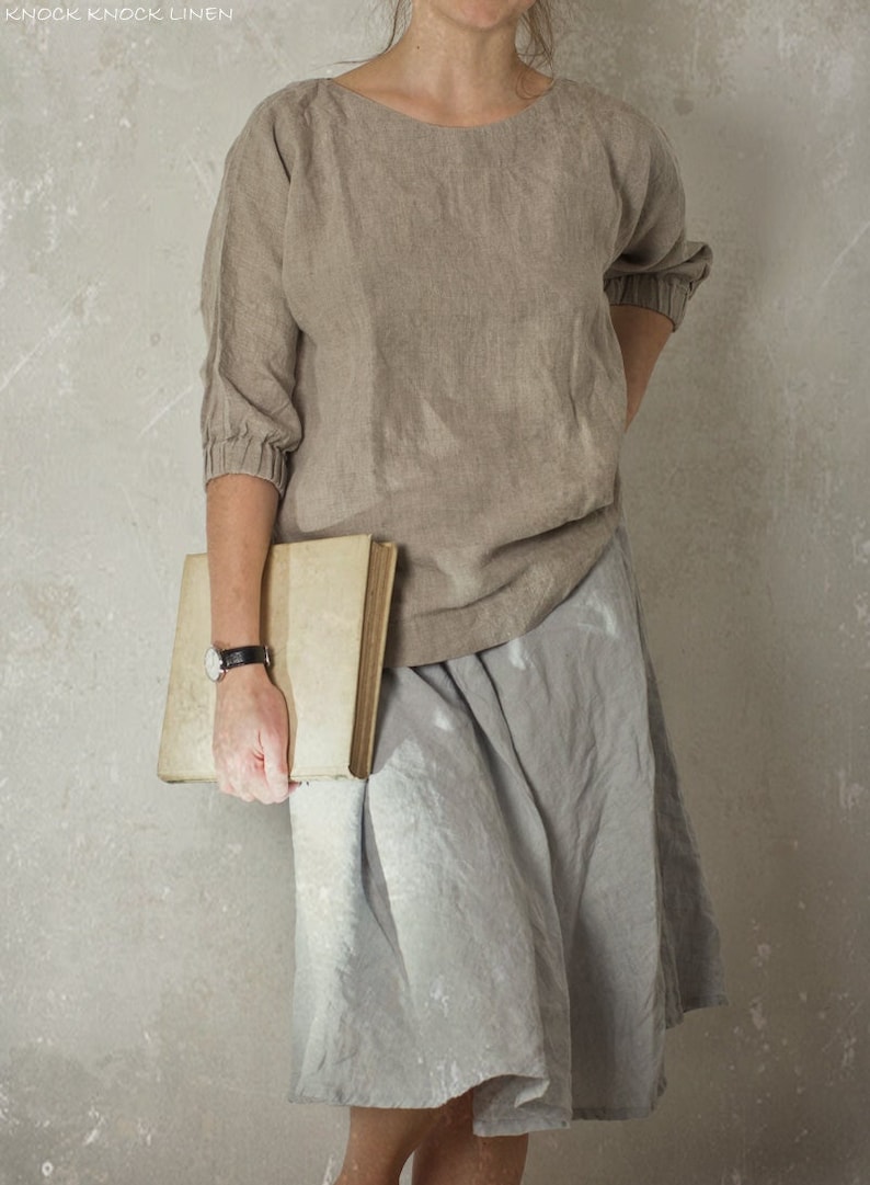 Woman wearing a loose linen blouse made from natural flax, featuring a boat neck and gathered cuffs, styled with a light gray skirt and black wristwatch, holding a book. Shown on model. Linen blouse, boat neck top.