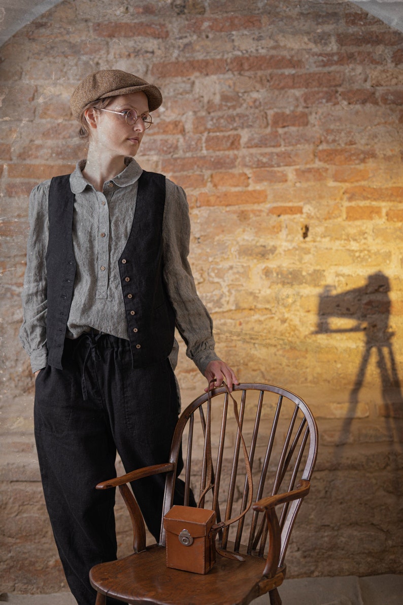 Linen pants in dark charcoal, shown on a model in a three-quarter front view, with a gathered elastic waistband, visible drawstring ties, loose legs, softly wrinkled fabric, and the model standing beside a wooden chair, Knock Knock Linen.