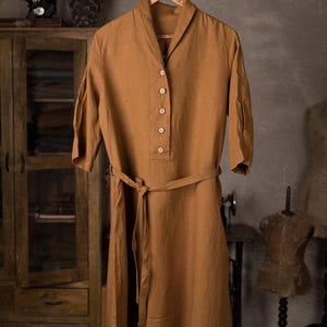 Warm brown linen belted dress displayed on hanger with rope loop, front full-length view, shawl-like collar, V opening, five buttons down the bodice, three-quarter sleeves, tied waist belt with long ends hanging; wooden cabinet and mannequin in background, knockknocklinen.