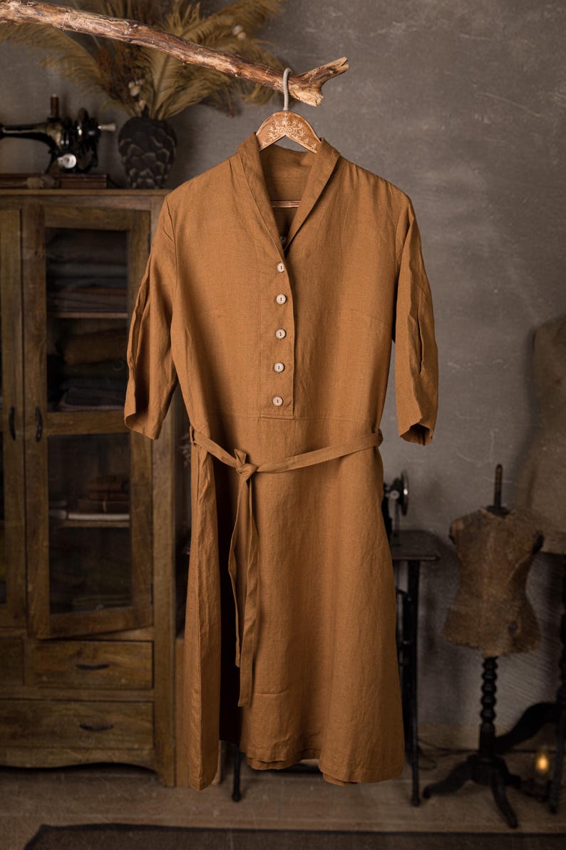 Warm brown linen belted dress displayed on hanger with rope loop, front full-length view, shawl-like collar, V opening, five buttons down the bodice, three-quarter sleeves, tied waist belt with long ends hanging; wooden cabinet and mannequin in background, knockknocklinen.