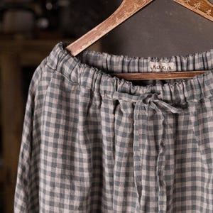 Gray and white gingham linen skirt shown full-length on an ornate wooden hanger, front view, ruched waistband with drawstring channel, smooth skirt panels with no visible pockets, wide hemline, cabinet and dress forms behind.