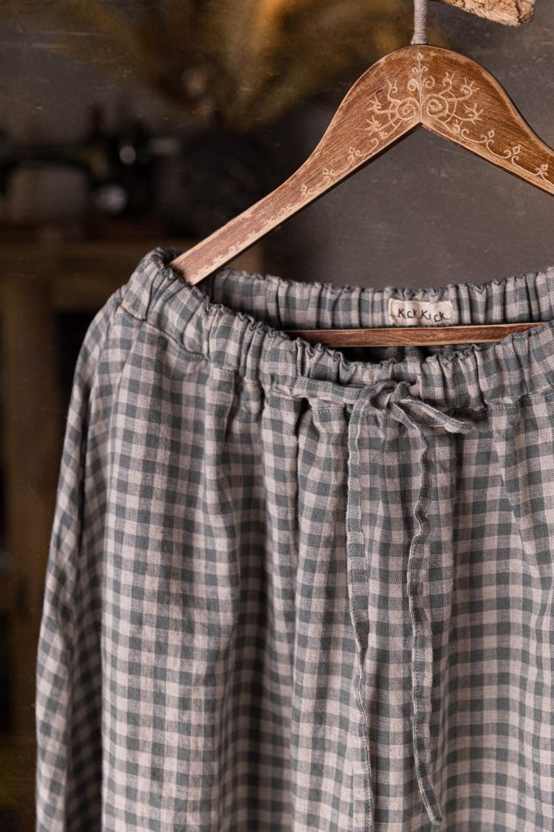 Gray and white gingham linen skirt shown full-length on an ornate wooden hanger, front view, ruched waistband with drawstring channel, smooth skirt panels with no visible pockets, wide hemline, cabinet and dress forms behind.