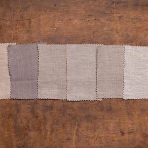 Linen fabric pieces in soft white, cream, beige, and taupe shades shown in a flat lay detail view, set in one row on worn wood, with pinked edges, mostly smooth plain weaves, and a textured light beige swatch on the right, Knock Knock Linen.