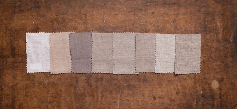 Linen fabric pieces in soft white, cream, beige, and taupe shades shown in a flat lay detail view, set in one row on worn wood, with pinked edges, mostly smooth plain weaves, and a textured light beige swatch on the right, Knock Knock Linen.
