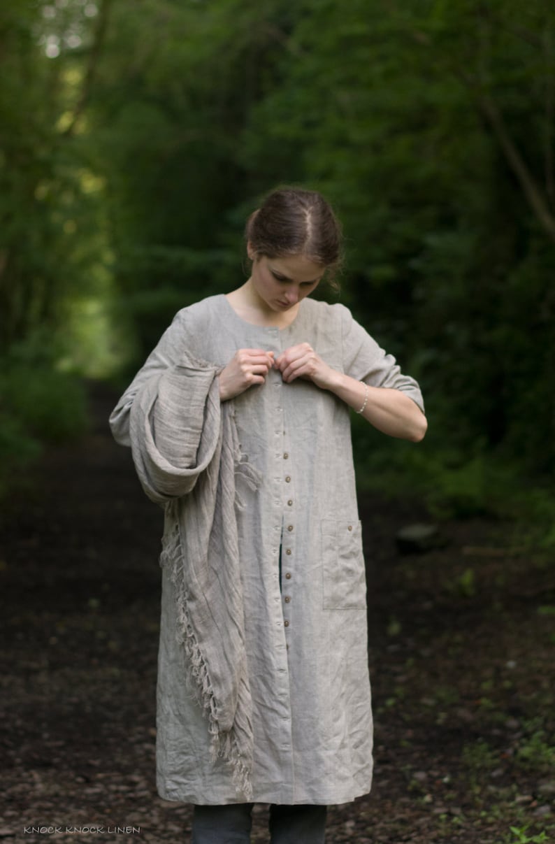 Natural flax linen dress coat with long sleeves, button front, and two patch pockets. The loose-fit buttoned dress is styled with a scarf, photographed outdoors against a forested backdrop.