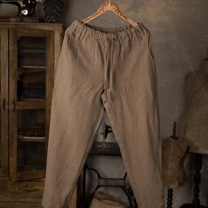 Gray-brown linen pants, shown hanging in a full-length front-facing photo, with an elastic waist, softly wrinkled fabric, rounded front pockets, a center front seam, and legs that narrow toward the hem, Knock Knock Linen.