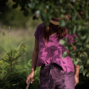 Woman wearing a raspberry-colored loose linen blouse with short kimono sleeves and a V-neck, paired with matching linen trousers and a wide-brimmed straw hat. She holds a small gardening trowel while standing among tall green plants outdoors.