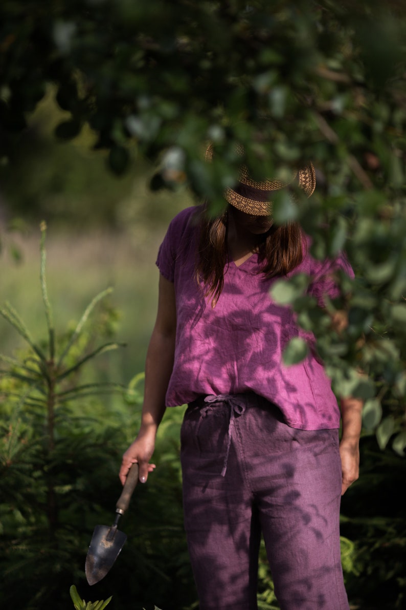 Woman wearing a raspberry-colored loose linen blouse with short kimono sleeves and a V-neck, paired with matching linen trousers and a wide-brimmed straw hat. She holds a small gardening trowel while standing among tall green plants outdoors.