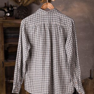 Full back view of a gray and white checked linen long-sleeve shirt on hanger, collar visible, gathered back panel under the yoke, buttoned cuffs, loose drape, photographed against a rustic cabinet and sewing studio backdrop, knockknocklinen.