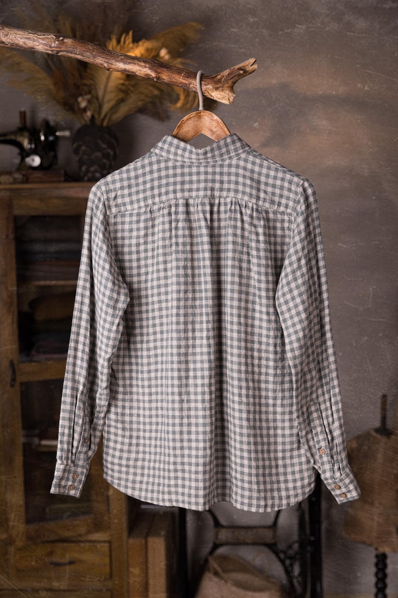 Full back view of a gray and white checked linen long-sleeve shirt on hanger, collar visible, gathered back panel under the yoke, buttoned cuffs, loose drape, photographed against a rustic cabinet and sewing studio backdrop, knockknocklinen.