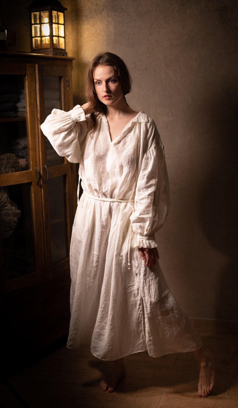 white linen smock dress with long puff sleeves, V-slit neckline, and thin waist tie belt, photographed barefoot beside a wooden cabinet.
