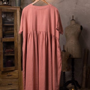 Dusty rose linen dress hanging on a carved wooden hanger from a rustic branch, front full-length view, round neckline, short sleeves, dropped waist seam with gathered skirt, loose drape, grey wall background.