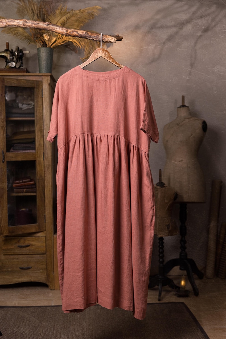 Dusty rose linen dress hanging on a carved wooden hanger from a rustic branch, front full-length view, round neckline, short sleeves, dropped waist seam with gathered skirt, loose drape, grey wall background.