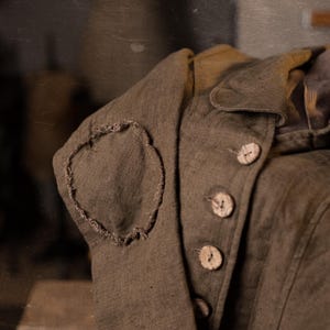 Detail crop of a muchroom  brown linen jacket, folded and photographed at an angle, showing the sleeve elbow patch with rough edges, the lapel fold, vertical seam lines, and a row of pale buttons on the front.