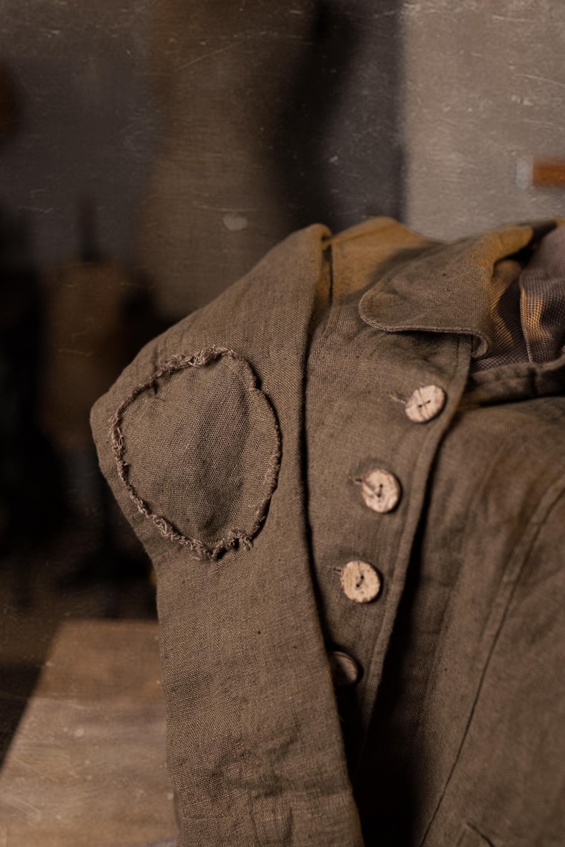 Detail crop of a muchroom  brown linen jacket, folded and photographed at an angle, showing the sleeve elbow patch with rough edges, the lapel fold, vertical seam lines, and a row of pale buttons on the front.