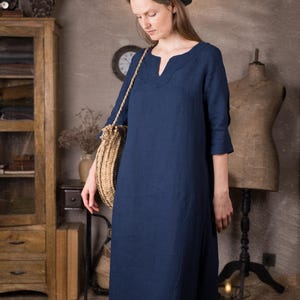 Front-side view of a model in a long dark blue linen summer dress with 3/4 sleeves and relaxed straight silhouette, standing on a rug in a neutral room with a grey hat and straw crossbody bag.