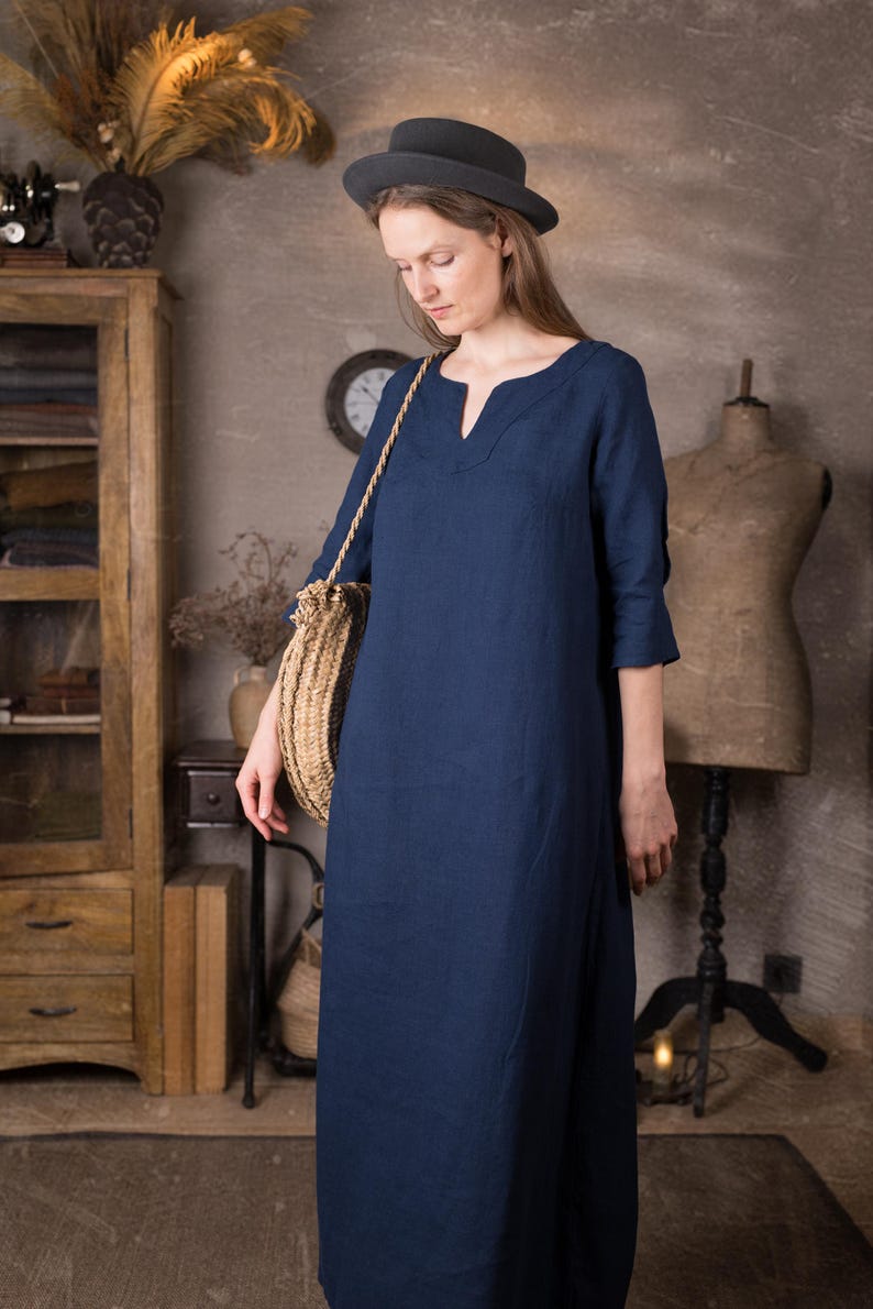 Front-side view of a model in a long dark blue linen summer dress with 3/4 sleeves and relaxed straight silhouette, standing on a rug in a neutral room with a grey hat and straw crossbody bag.
