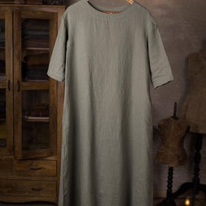 linen dress displayed on hanger from a branch, front view full-length crop, wide round neck, short sleeves with simple hems, smooth unbroken front panel, straight hemline; grey wall studio background.