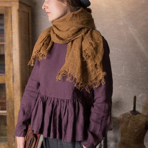 Side view of a woman indoors wearing a dark plum long sleeve linen peplum blouse with gathered hem, styled with a large brown scarf, grey trousers and a black hat.