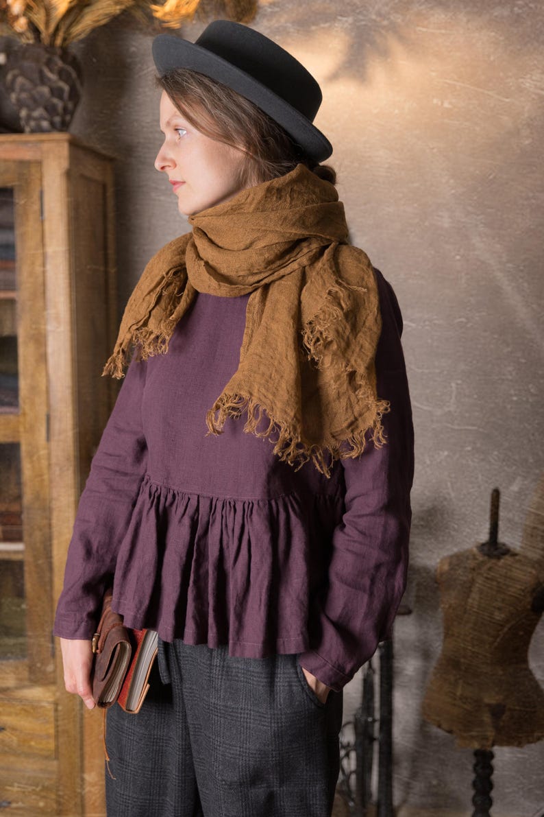 Side view of a woman indoors wearing a dark plum long sleeve linen peplum blouse with gathered hem, styled with a large brown scarf, grey trousers and a black hat.