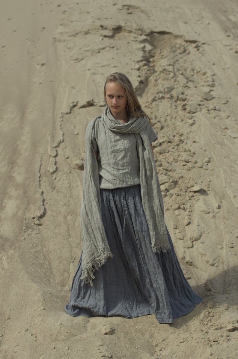 Model outdoors wearing a frost gray linen skirt with drawstring waist, elastic waistband, and side pockets; floor-length, loose fit in cottage-style. Paired with light gray linen top and scarf. Keywords: maxi linen skirt, peasant skirt.