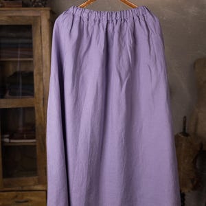 Lavender linen maxi skirt shown full-length on an ornate wooden hanger, front view, elasticated gathered waist, drawstring channel with hanging ties, wide A-line shape, curved hemline, cabinet and mannequin forms in the background.