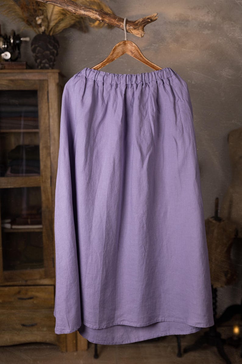 Lavender linen maxi skirt shown full-length on an ornate wooden hanger, front view, elasticated gathered waist, drawstring channel with hanging ties, wide A-line shape, curved hemline, cabinet and mannequin forms in the background.