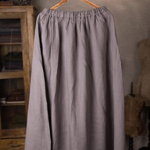 Gray linen skirt shown full-length on a hanger, front view, elasticated gathered waist with drawstring channel, dangling ties, wide A-line shape, plain front with no visible pockets, wooden cabinet and mannequins in background.