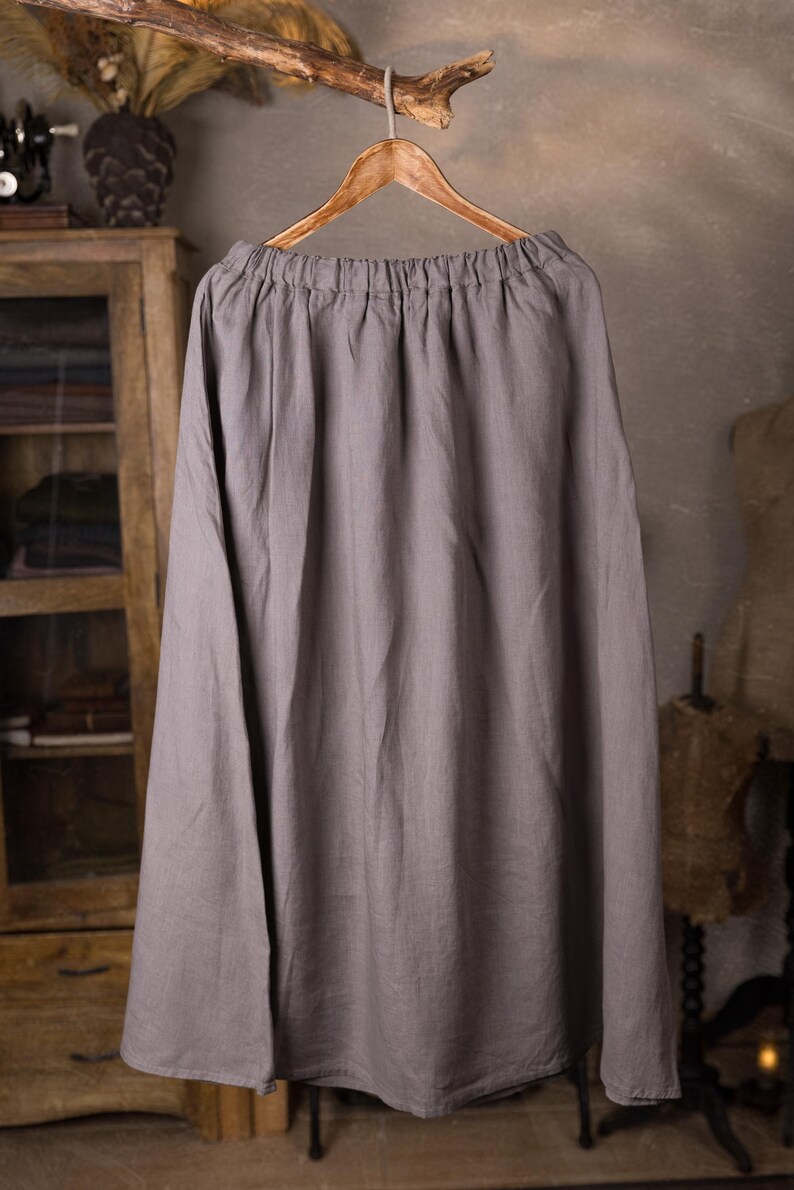 Gray linen skirt shown full-length on a hanger, front view, elasticated gathered waist with drawstring channel, dangling ties, wide A-line shape, plain front with no visible pockets, wooden cabinet and mannequins in background.